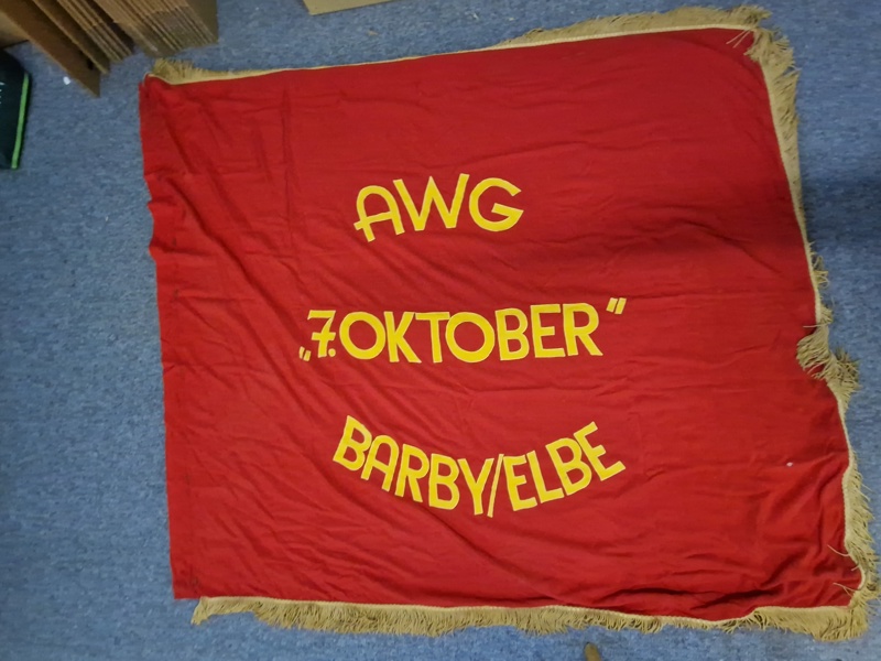 fane med teksten "AWG", "7. OKTOBER" og "BARBY/ELBE".
AWG: står for "Arbeiterwohnungsbaugenossenschaft" (Arbejderboligbyggeriets Andelsforening), som var en type boligforening i DDR.