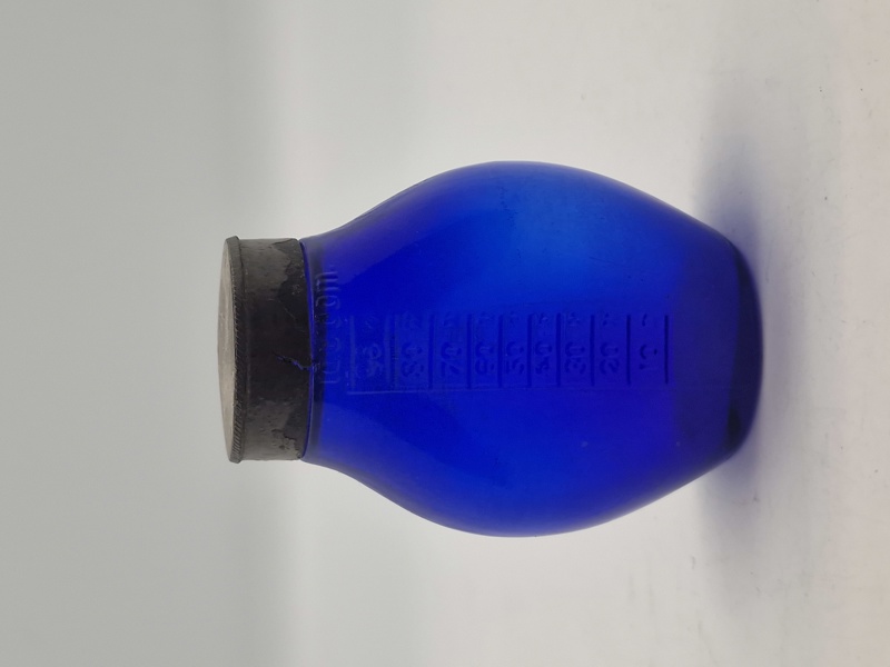 Blå spytflaske af glas med metalkapsel og gradueret skala i kubikcentimeter, som blev brugt til at opsamle spyt fra patienter med tuberkulose, især i perioden 1871-1920